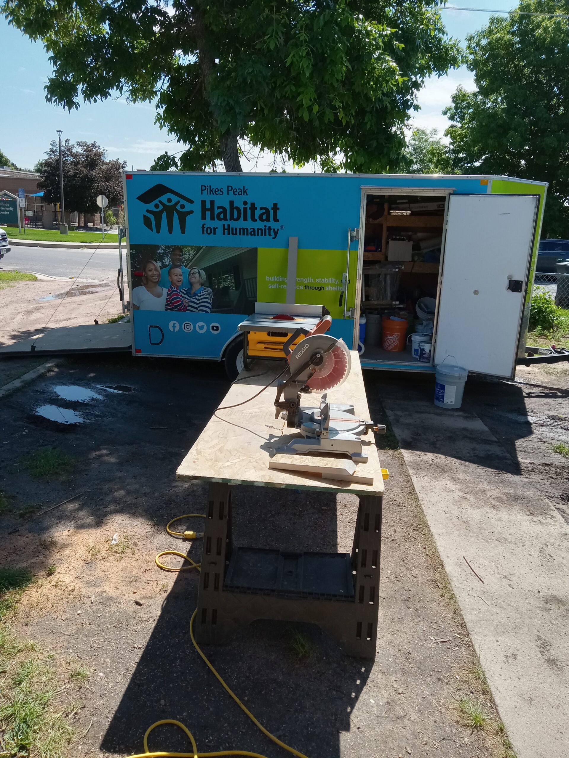 Home repair program trailer in yard with saw and table set up in front