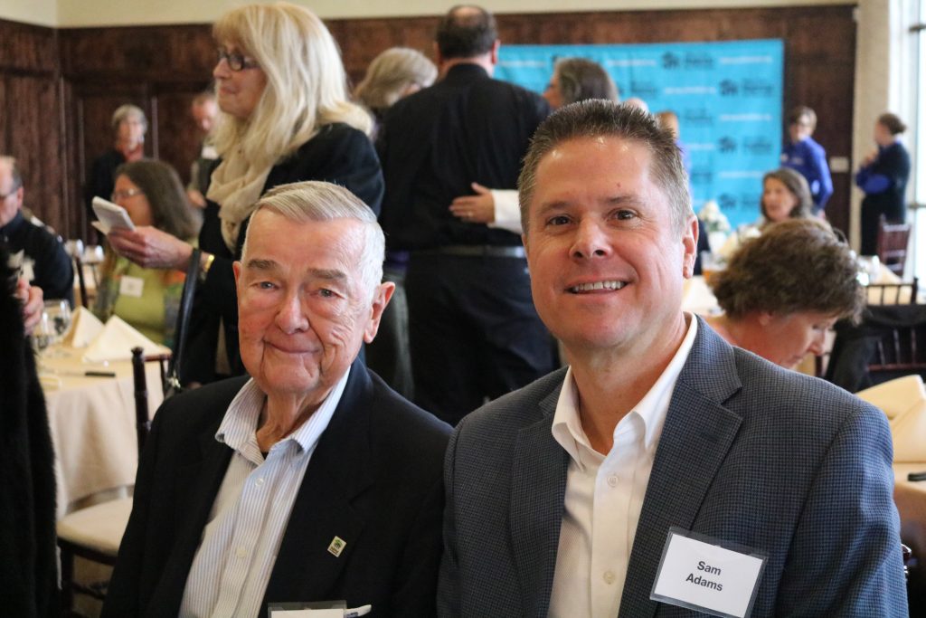 Two gentlemen sit side-by-side smiling at the camera. The man on the right has a name tag that reads "Sam Adams." In the background are many people talking to one another at an event along with tables covered in linens and a Habitat for Humanity photo wall.