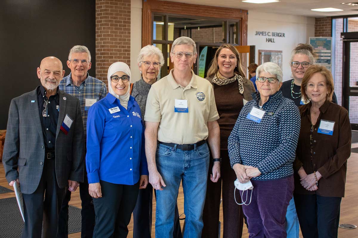A group of diverse individuals, including men and women of various ages, are posed standing in two rows and smiling at the camera. Everyone is wearing a "Committee Member" ribbon, with one woman wearing a shirt that says "Pikes Peak Habitat for Humanity Team Member."