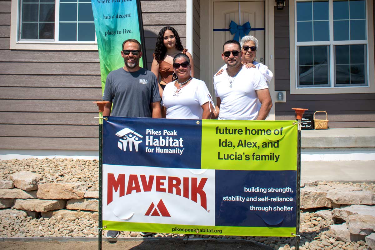 A family stands in front of a banner that reads "Pikes Peak Habitat for Humanity: Future home of Ida, Alex, and Lucia's Family." A "Maverik" logo is placed at the bottom of the banner.