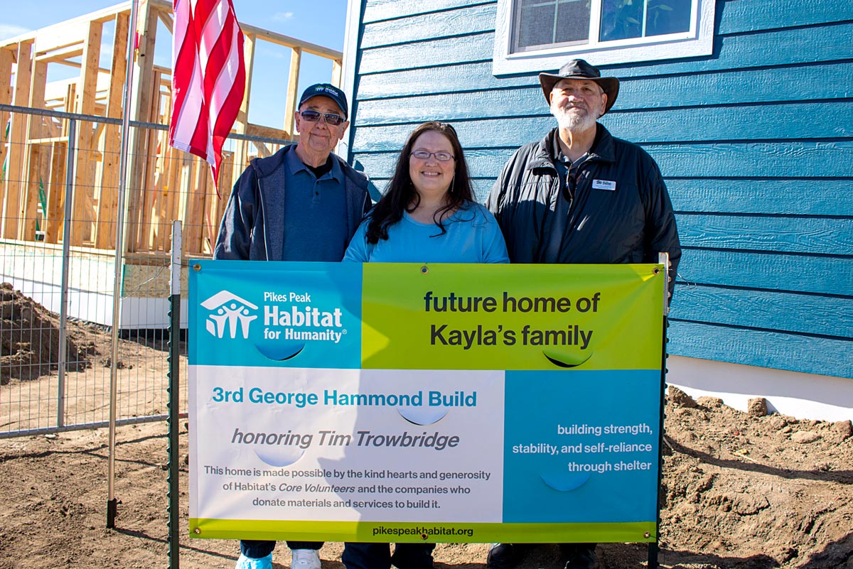 Two men and a woman stand in front of a sign that reads "3rd George Hammond Build" with a Pikes Peak Habitat for Humanity logo