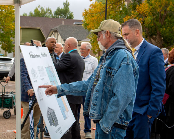A man in a Canadian Tuxedo points at a sign with a rendering of a house and a Habitat for Humanity logo
