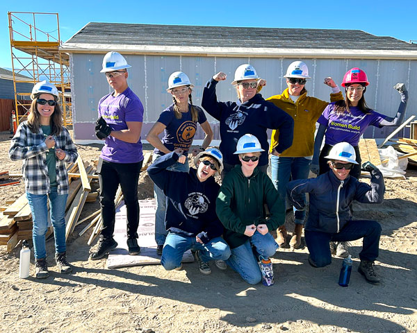 A group of volunteers on a construction site wearing hard hats flex their muscles